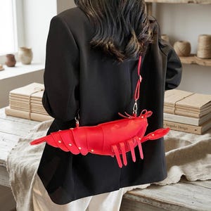 May include: A bright red crossbody bag shaped like a shrimp or lobster, featuring segmented body panels, small gold-toned studs, and a thin adjustable shoulder strap. The bag has a curved tail section and multiple dangling leg-like appendages.