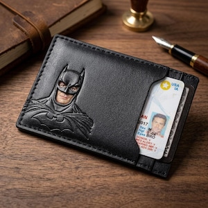 May include: A black leather card holder featuring an embossed Batman emblem on the front. The wallet contains a visible identification card with the text USA IA, 2017, and donor information. The design includes a single slot for cards.