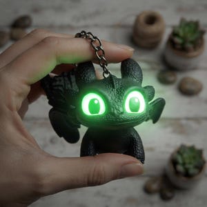 May include: A black dragon keychain featuring a textured, scaled surface and large, glowing green eyes. The figure has small wings, pointed ears, and a metal chain attached to the top of its head. The eyes emit a bright, neon green light.