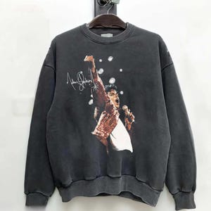 May include: Dark gray sweatshirt featuring a graphic of a performer with a microphone, wearing a red jacket. The sweatshirt has the performer's signature and a vintage wash. The design is centered on the front of the crew neck sweatshirt.