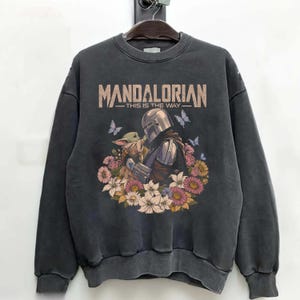 K&ouml;nnte beinhalten: Dunkelgraues Sweatshirt mit einer Grafik von The Mandalorian und Grogu, umgeben von Blumen und Schmetterlingen. Der Text "MANDALORIAN - THIS IS THE WAY" ist oben in Beige aufgedruckt.