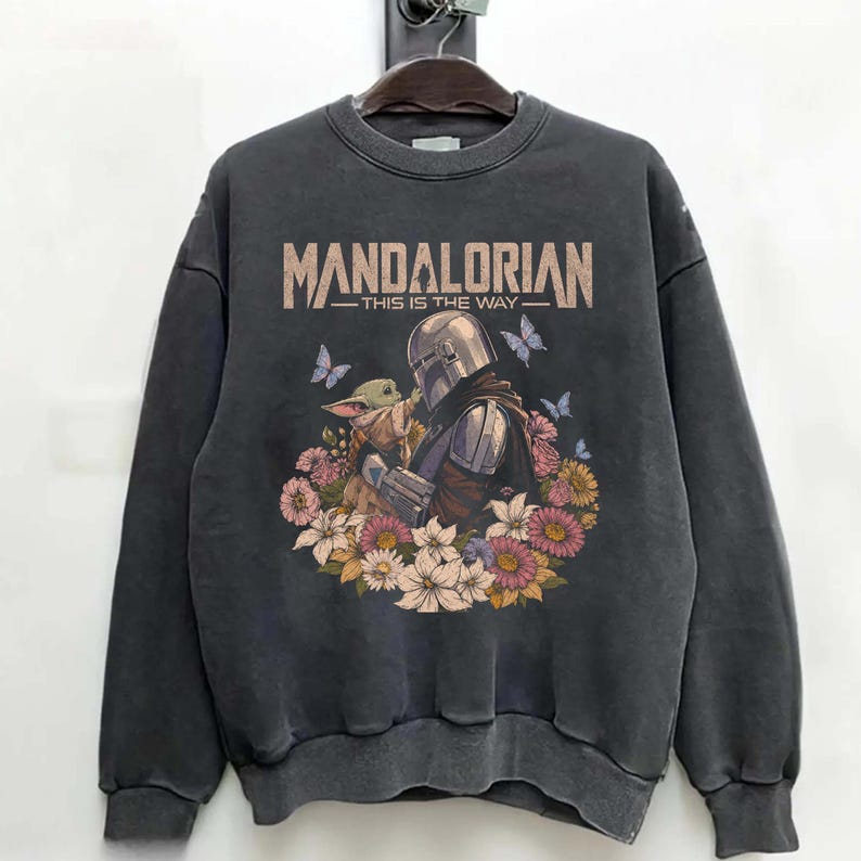 K&ouml;nnte beinhalten: Dunkelgraues Sweatshirt mit einer Grafik von The Mandalorian und Grogu, umgeben von Blumen und Schmetterlingen. Der Text "MANDALORIAN - THIS IS THE WAY" ist oben in Beige aufgedruckt.