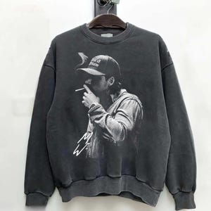 Op de afbeelding: Donkergrijze sweatshirt met een zwart-witte afbeelding van een persoon die een sigaret rookt en een baseballpet draagt. Op de pet staat de tekst "Field & Stream". Onder de afbeelding staat een handtekening.