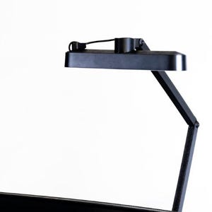 Puede incluir: Una lámpara de escritorio negra y ajustable con un cabezal de luz rectangular y un brazo flexible. La lámpara es de metal y tiene un diseño moderno. La lámpara está diseñada para montarse en un escritorio o mesa.