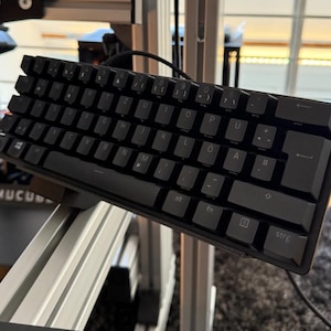 May include: A compact black mechanical keyboard with a QWERTZ layout, featuring dark keys with white lettering. The keyboard includes a Windows key, a function key, and a strg key, connected by a black cable and mounted on a silver metal frame.