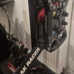 Puede incluir: Un volante negro de simulador de carreras con múltiples botones y diales. El volante está montado en un marco negro con el texto "TRAK RACER" en blanco. El volante tiene el texto "SIMAGIC" en rojo y blanco.