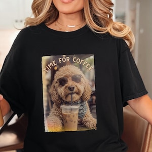 May include: A black short-sleeved t-shirt featuring a graphic print of a light brown curly-haired dog wearing sunglasses and a patterned bandana. The dog is drinking from a clear plastic cup with a straw. The text TIME FOR COFFEE is printed above.