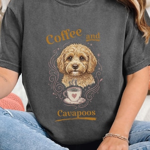 May include: A charcoal grey short-sleeve t-shirt featuring a graphic of a golden-brown Cavapoo dog above a steaming coffee cup with a heart design. The text "Coffee and Cavapoos" is printed in gold lettering above and below the dog illustration.