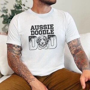 May include: A white short-sleeved t-shirt featuring the black text "AUSSIE DOODLE DAD" printed on the chest. The letter "O" in the word "DOD" is replaced by a detailed black and white illustration of an Australian Doodle dog face.