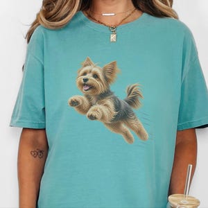 May include: A teal short-sleeved t-shirt featuring a graphic illustration of a small, tan and black Yorkshire Terrier dog in mid-air, appearing to jump or run. The dog has pointed ears, a fluffy coat, and an open mouth with a visible tongue.