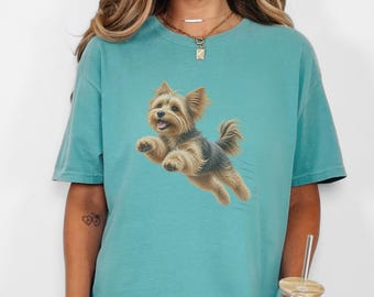 May include: A teal short-sleeved t-shirt featuring a graphic illustration of a small, tan and black Yorkshire Terrier dog in mid-air, appearing to jump or run. The dog has pointed ears, a fluffy coat, and an open mouth with a visible tongue.