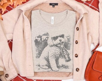May include: A light heather gray crew neck t-shirt featuring a dark gray graphic print of a fluffy dog wearing sunglasses and holding a beer bottle labeled with three X's in its mouth. The shirt is layered under a light beige button-down jacket.