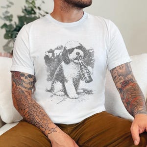 May include: A white short-sleeved t-shirt featuring a gray graphic print of a dog wearing sunglasses and holding a beer bottle labeled with XXX. The design is centered on the chest of the shirt, showcasing a detailed illustration of the canine.