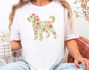 Puede incluir: Una camiseta blanca de manga corta con un gráfico central de la silueta de un perro compuesta enteramente por varias flores coloridas y hojas verdes. El diseño floral incluye tonos de rosa, naranja, amarillo y flores blancas con follaje verde.