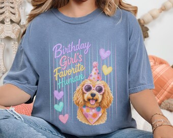 May include: A blue t-shirt featuring a graphic of a golden-colored dog wearing pink sunglasses, a polka-dot party hat, and a matching bandana. The text reads "Birthday Girl's Favorite Human" in purple and yellow, surrounded by colorful hearts.