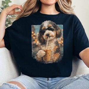 Könnte beinhalten: Ein schwarzes T-Shirt mit einem Grafikdruck eines flauschigen Hundes, der eine schwarze Sonnenbrille trägt und einen Eiskaffee aus einem durchsichtigen Plastikbecher mit Strohhalm trinkt. Der Hund hat braunes, schwarzes und weißes Fell.