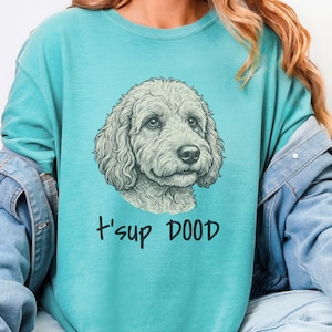 Puede incluir: Una sudadera de cuello redondo color verde azulado con una ilustración detallada de líneas de la cara de un perro de pelo rizado en el centro. Debajo de la ilustración del perro, el texto "t'sup DOOD" está impreso en una fuente negra y negrita.