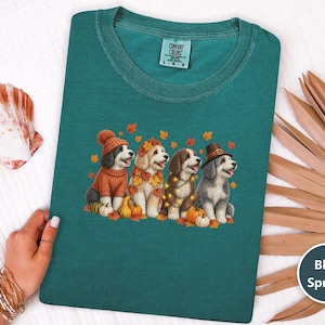 Pode incluir: Uma t-shirt de cor azul abeto com um gráfico de quatro cães a usar acessórios de outono, incluindo um gorro de malha, uma coroa de folhas, luzes de corda e um chapéu de peregrino, rodeados por pequenas abóboras e folhas de outono.