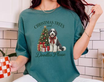 May include: A dark teal t-shirt featuring a graphic of a black and white doodle dog wearing a red plaid bow, sitting next to a decorated Christmas tree and wrapped gifts. The text reads, "CHRISTMAS TREES and Doodles Please."
