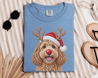 May include: A light blue crew neck t-shirt featuring a graphic of a curly-haired brown dog wearing a red and white Santa hat, brown reindeer antlers, and a bright red nose. The shirt has a Comfort Colors brand label at the inner neckline.
