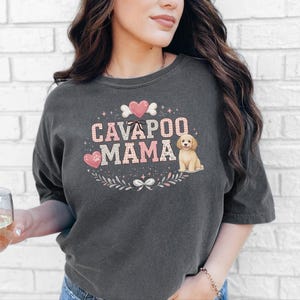 May include: A charcoal grey short-sleeve t-shirt featuring a graphic print with the text CAVAPOO MAMA in pink patterned letters. The design includes a small illustration of a light brown dog, pink hearts, a bone, and floral accents.