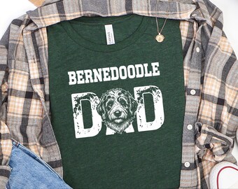 Peut inclure: Un t-shirt à manches courtes vert foncé avec le texte blanc "BERNEDOODLE DAD" et une illustration détaillée d'une tête de chien Bernedoodle remplaçant la lettre A. Le t-shirt est superposé sous une chemise à carreaux beige, noire et blanche.