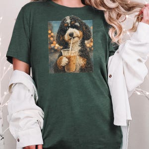 May include: A dark forest green short-sleeve t-shirt featuring a graphic print of a curly-haired dog wearing black sunglasses and holding a clear plastic cup with a straw filled with an iced coffee beverage against a blurred light background.