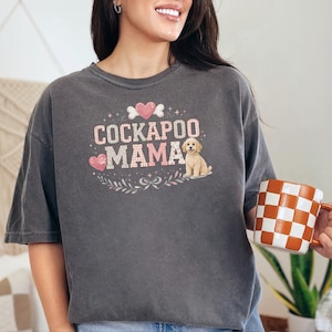 May include: A charcoal gray short-sleeve t-shirt featuring a graphic print that reads COCKAPOO MAMA in pink and white checkered lettering, accompanied by a small illustration of a light-colored puppy, pink hearts, and decorative floral elements.