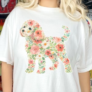Può includere: Una t-shirt bianca a maniche corte con una stampa grafica raffigurante la sagoma di un cane composta interamente da vari fiori rosa, rossi e bianchi insieme a foglie verdi. Il disegno floreale del cane è centrato sulla parte anteriore.