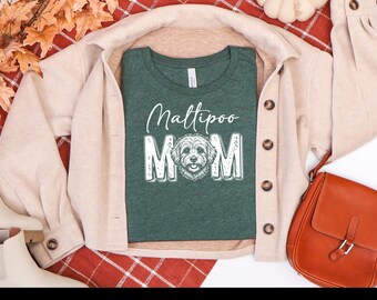 May include: A dark green short-sleeved t-shirt featuring the white text Maltipoo MOM with a detailed line-art illustration of a Maltipoo dog face replacing the letter O. The shirt is layered under a beige button-down jacket.
