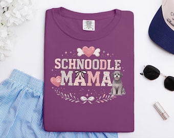 May include: A plum-colored t-shirt featuring the text "SCHNOODLE MAMA" in a pink and white patterned font. The design includes a graphic of a gray Schnoodle dog, pink hearts, a black bow, and white floral accents surrounding the text.