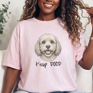 May include: A light pink short-sleeved t-shirt featuring a graphic illustration of a curly-haired dog face centred on the chest. Below the dog illustration, the text t'sup DOOD is printed in a casual, dark-coloured handwritten font.
