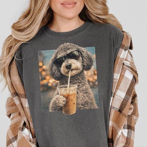 Könnte beinhalten: Ein anthrazitfarbenes T-Shirt mit Rundhalsausschnitt und einem Grafikdruck eines lockigen Hundes, der eine schwarze Sonnenbrille trägt und einen Plastikbecher mit Strohhalm vor einem Hintergrund aus unscharfen Lichtern hält.