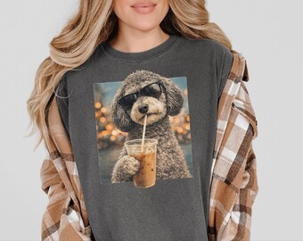 May include: A charcoal gray crew neck t-shirt featuring a graphic print of a curly-haired dog wearing black sunglasses and holding a plastic cup with a straw. The dog is positioned against a blurred background of warm, glowing lights.