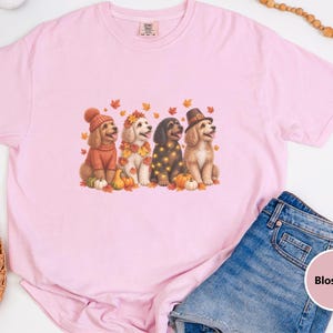 Puede incluir: Una camiseta rosa claro con un gráfico de cuatro perros sentados entre hojas de otoño y calabazas. Los perros llevan accesorios de temporada que incluyen un gorro de punto, una corona de hojas, luces decorativas y un sombrero de peregrino.