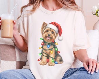 May include: A cream-colored t-shirt featuring a graphic illustration of a Yorkshire Terrier wearing a red and white Santa hat. The dog is wrapped in a string of colorful, glowing Christmas lights in red, green, blue, and yellow hues.