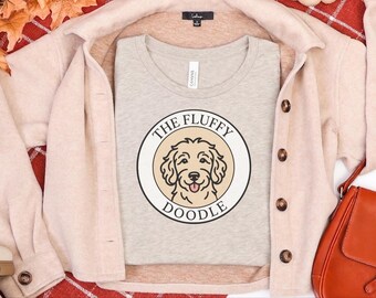 Peut inclure: Un t-shirt à col rond beige chiné avec un motif circulaire représentant un chien doodle beige et le texte THE FLUFFY DOODLE en noir. Le t-shirt est superposé sous une chemise beige clair boutonnée avec des boutons marron.