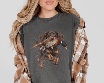 May include: A charcoal gray crew neck t-shirt featuring a graphic illustration of a black and tan dachshund dog running while carrying a pheasant in its mouth. The dog is depicted with detailed fur textures and the pheasant has colorful plumage.