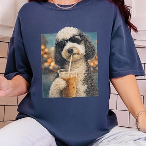 May include: A navy blue short-sleeve t-shirt featuring a graphic print of a fluffy black and white dog wearing black sunglasses and drinking an iced coffee from a clear plastic cup with a straw against a blurred bokeh background.