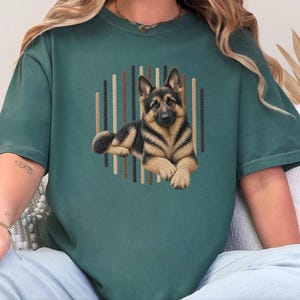 Può includere: Una t-shirt a girocollo verde scuro con la grafica di un pastore tedesco sdraiato. Il cane è centrato davanti a uno sfondo circolare composto da strisce verticali nelle tonalità del marrone, del beige e del nero.