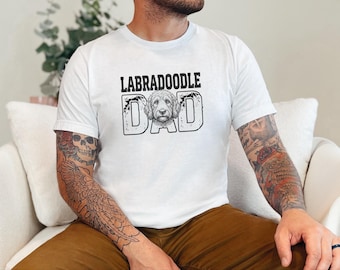 May include: A white short-sleeved t-shirt featuring a black graphic print on the chest. The design includes the text LABRADOODLE DAD in a bold font, with the letter O replaced by a detailed black and white illustration of a dog face.