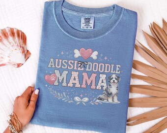 May include: A blue crew neck t-shirt featuring a graphic print with the text "AUSSIEDOODLE MAMA" in a block font. The design includes a pink heart, a small black bow, and a seated merle Aussiedoodle puppy surrounded by floral accents.