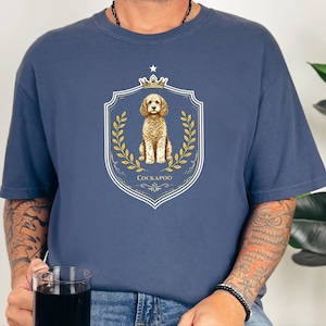 May include: A slate blue short-sleeved t-shirt featuring a central graphic of a golden-coloured Cockapoo dog sitting inside a white shield emblem. The design includes a small crown at the top, golden laurel branches on the sides, and the text COCKAPOO.