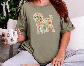 May include: An olive green t-shirt featuring a graphic of a dog silhouette filled with a colorful floral pattern of pink, orange, and white flowers. The shirt is paired with a white mug decorated with black tree outlines and a candy cane.