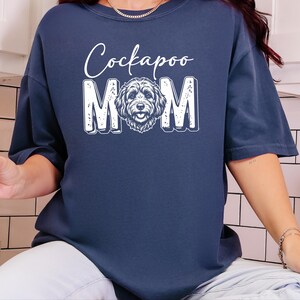 May include: A navy blue t-shirt featuring a white graphic print that reads "Cockapoo MOM" with the letter "O" replaced by a detailed illustration of a Cockapoo dog face. The design has a distressed texture and is centered on the chest.