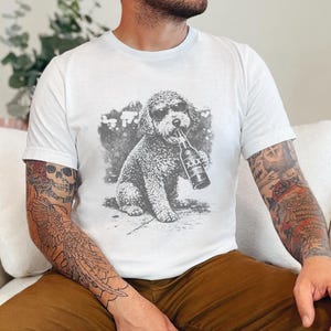 May include: A white short-sleeved t-shirt featuring a black and white graphic print of a curly-haired dog wearing sunglasses and holding a beer bottle labeled with three X symbols in its mouth. The design is centered on the chest area.