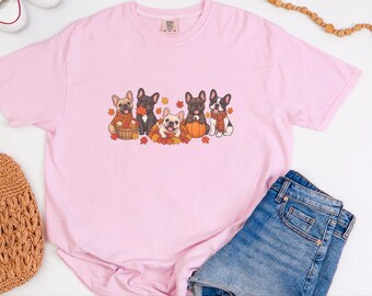 May include: A light pink short-sleeve t-shirt featuring a graphic of five French bulldogs surrounded by autumn leaves, a basket, and a pumpkin. The dogs are depicted in various colors including fawn, black, and pied, some wearing scarves and sweaters.