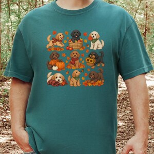 May include: A teal short-sleeve t-shirt featuring a graphic print of nine cartoon puppies in various poses surrounded by autumn leaves, pumpkins, sunflowers, and apples. The puppies are depicted in shades of gold, brown, and black.