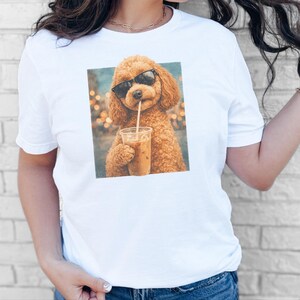 May include: A white short-sleeved t-shirt featuring a square graphic print of a fluffy brown poodle wearing black sunglasses and drinking an iced coffee from a clear plastic cup with a straw against a blurred background of warm golden lights.