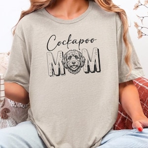 Puede incluir: Una camiseta beige de manga corta con el texto negro "Cockapoo" sobre la palabra "MOM" en letras de bloque, con una ilustración detallada en blanco y negro de la cara de un perro Cockapoo que reemplaza la letra "O" en la palabra "MOM".